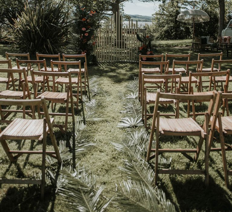 Boho outdoor wedding ceremony with wooden chairs, white wooden post and tropical flower installation and carpet of palm leaves for birds of paradise wedding inspiration