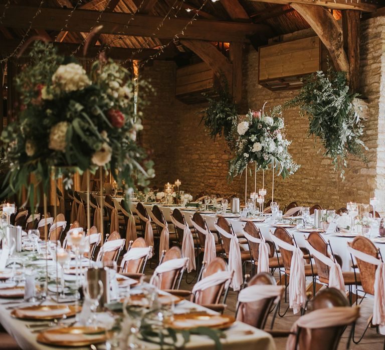 Brown leather chairs with blush pink fabric decor at long wooden breakfast tables with flower bombs and strung fairy lights inside stone room at Tythe Barn wedding with barn wedding flowers