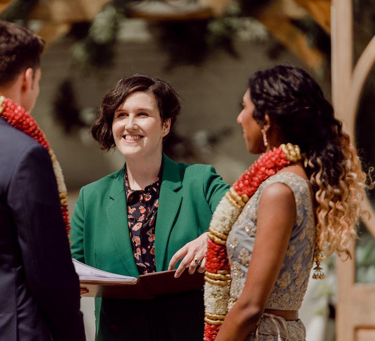 Celebrant in a green suit performing the multicultural outdoor wedding ceremony with Indian Bride and groom wearing flower garlands