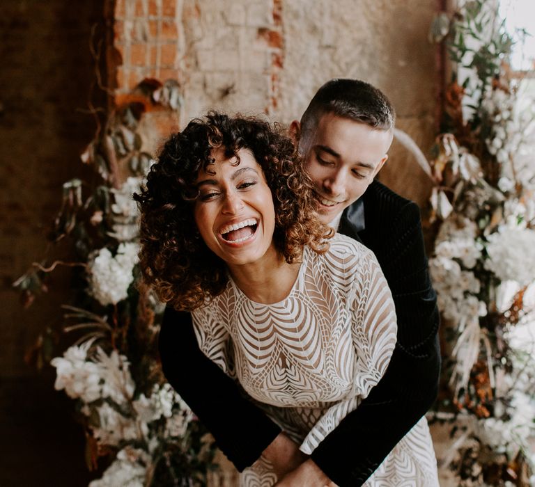 Groom embracing his laughing bride with naturally curly hair wearing a swirling patterned boho wedding dress 