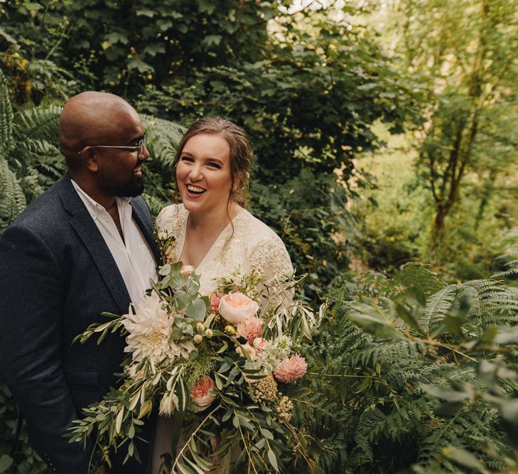 Black groom and white bride stand next to each other smiling, with bride holding wedding bouquet