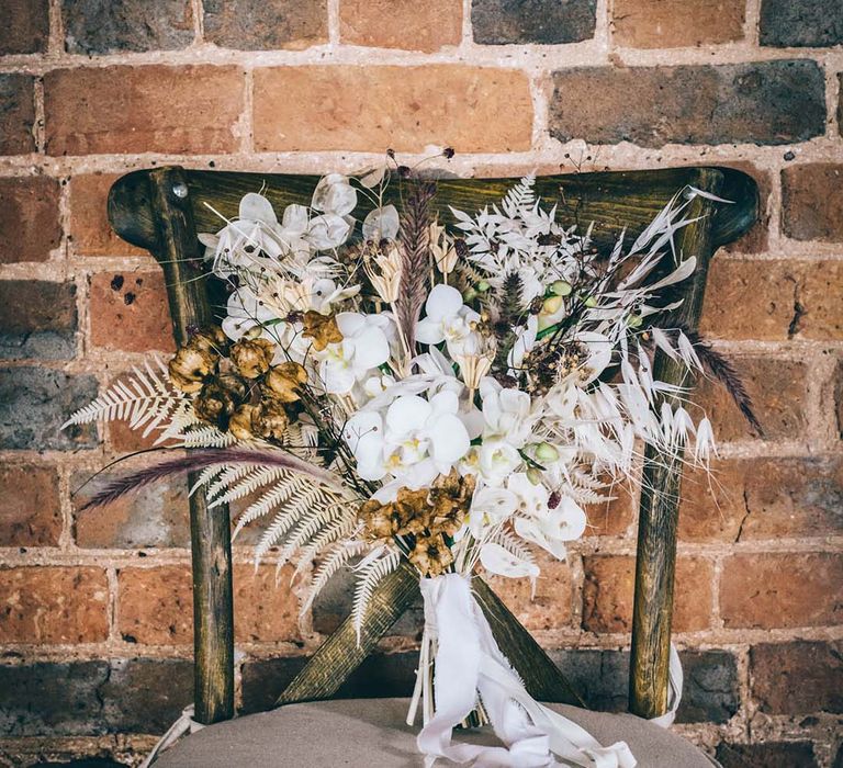 Fresh and dried flower wedding bouquet with white orchids resting on a chair 