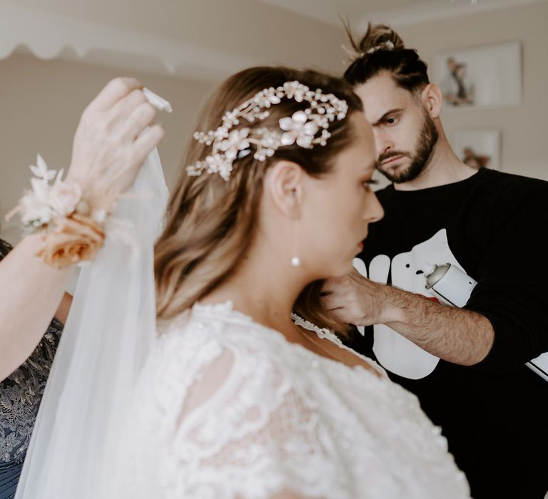 Bride in homemade lace wedding dress and floral and pearl hairpiece has her veil put on by wedding guest in blue dress before DIY garden wedding in Bedfordshire