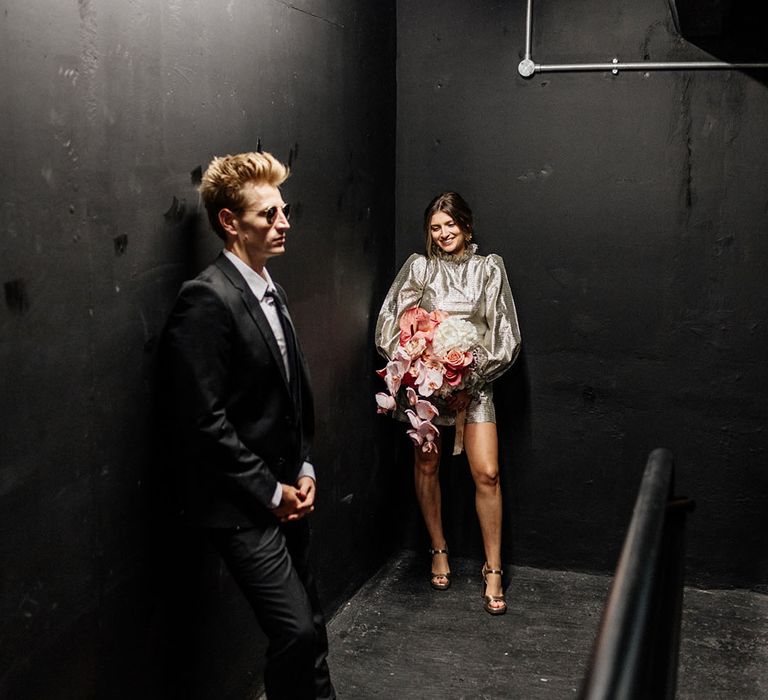 Bride in short gold wedding dress and groom in black suit with sunglasses on standing in dark surroundings industrial wedding