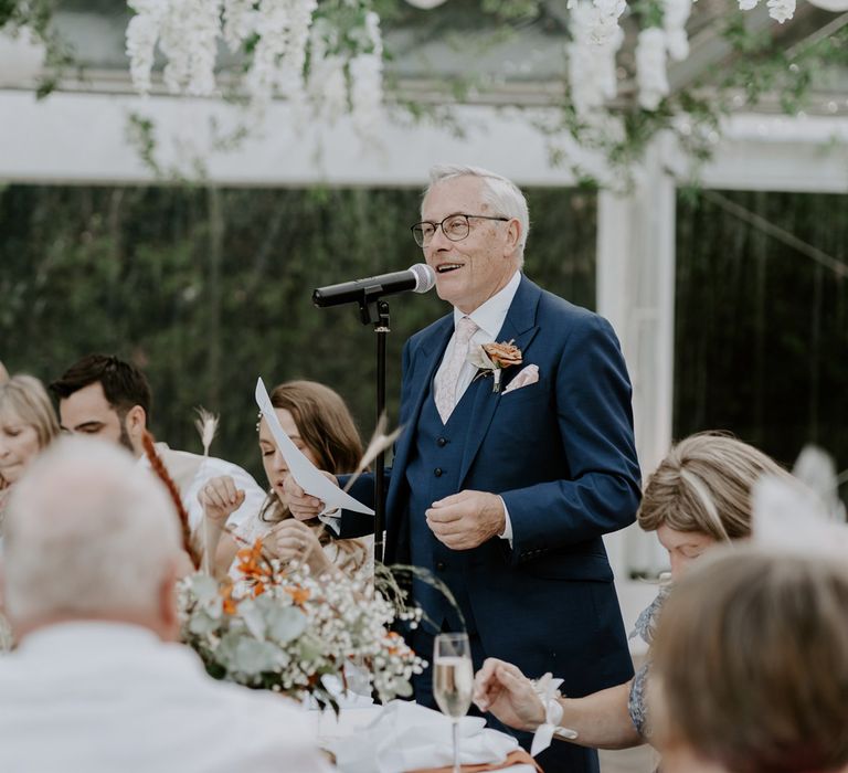 Man in blue three piece suit with burnt orange buttonhole makes speech during marquee wedding reception inn Bedfordshire 