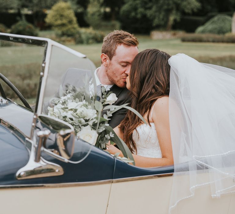 Bride in Pronovias wedding dress and veil holding white and green bridal bouquet kisses groom in grey suit as they sit in vintage car at Notley Abbey wedding