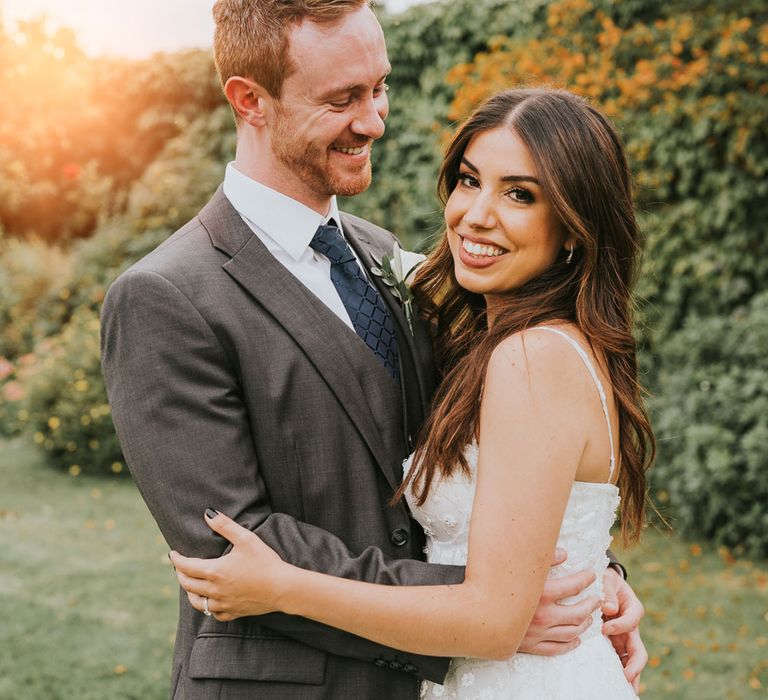 Groom in grey three piece suit, blue tie and white rose buttonhole smiles whilst looking at bride in white cami strap Pronovias wedding dress in the grounds of Notley Abbey in Buckinghamshire