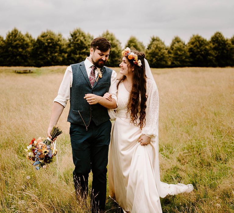 Bride & groom embrace in field as they walk along together and groom holds bridal bouquet