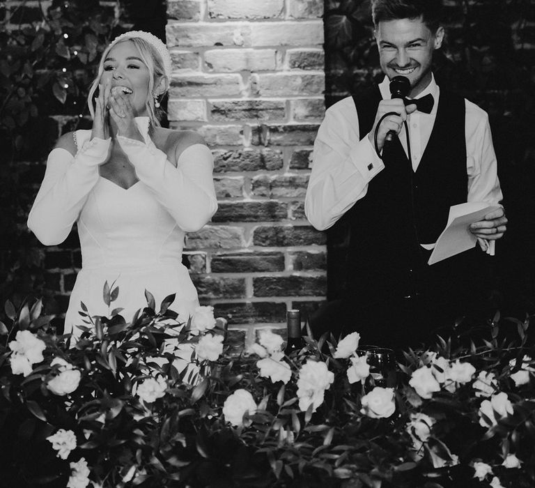 Black and white picture of the bride and groom standing during the wedding reception speeches. The bride wears a pearl headband 