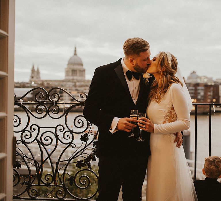 Groom in a tuxedo and bride in a long sleeve wedding dress kissing on a balcony with the London skyline in the background 