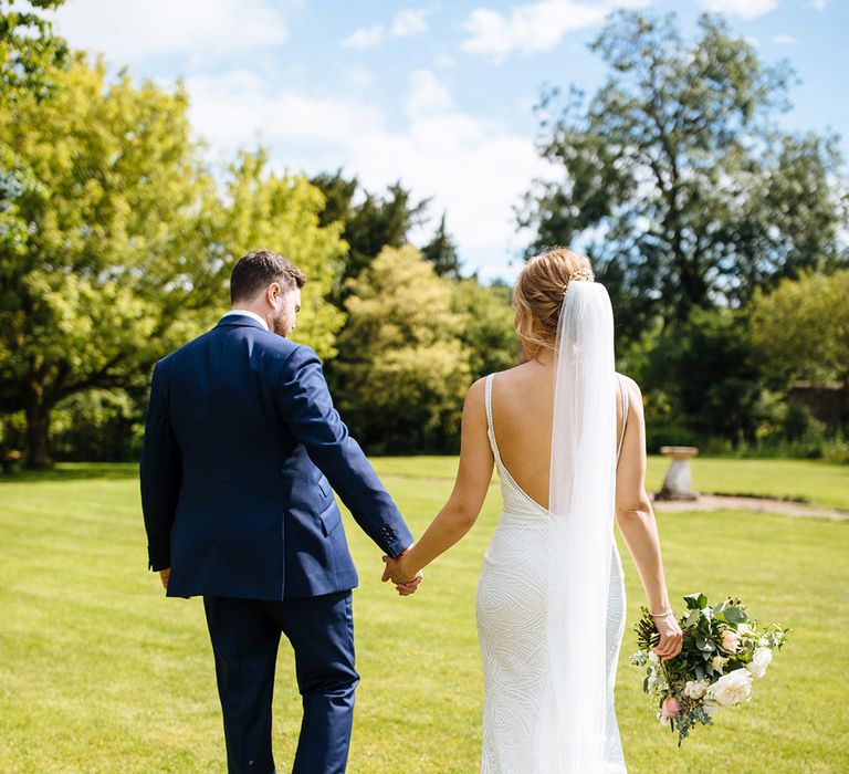 Groom in a navy blue suit holding hands with his bride in a white beaded wedding dress with low back and cathedral length veil 