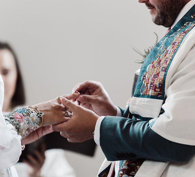 Groom in white and green tallit places ring on brides finger as she wears mesh sleeve wedding dress with embroidered cuff sleeves 