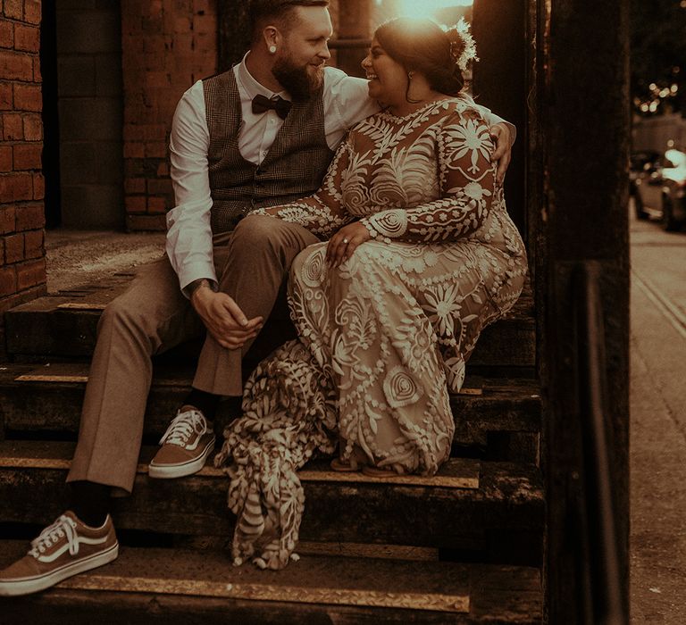 Golden house wedding portrait with groom in a tweed waistcoat and bride in a boho lace wedding dress sitting on the steps outside their Victoria Warehouse wedding venue 