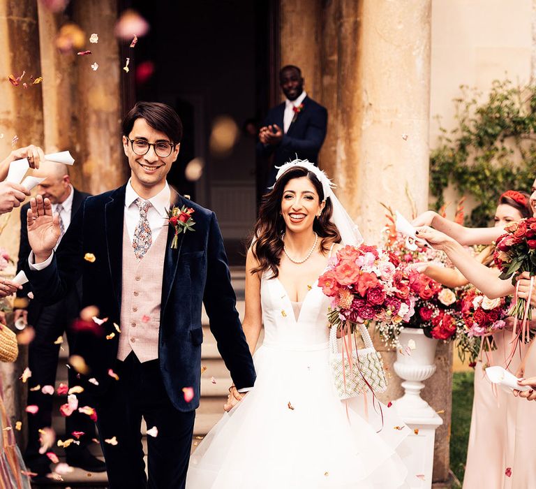 Bride in feathered headband and layered tulle wedding dress holding beaded bag and pink, white and red bridal bouquet walks through petal confetti with groom in dark suit with pink waistcoat