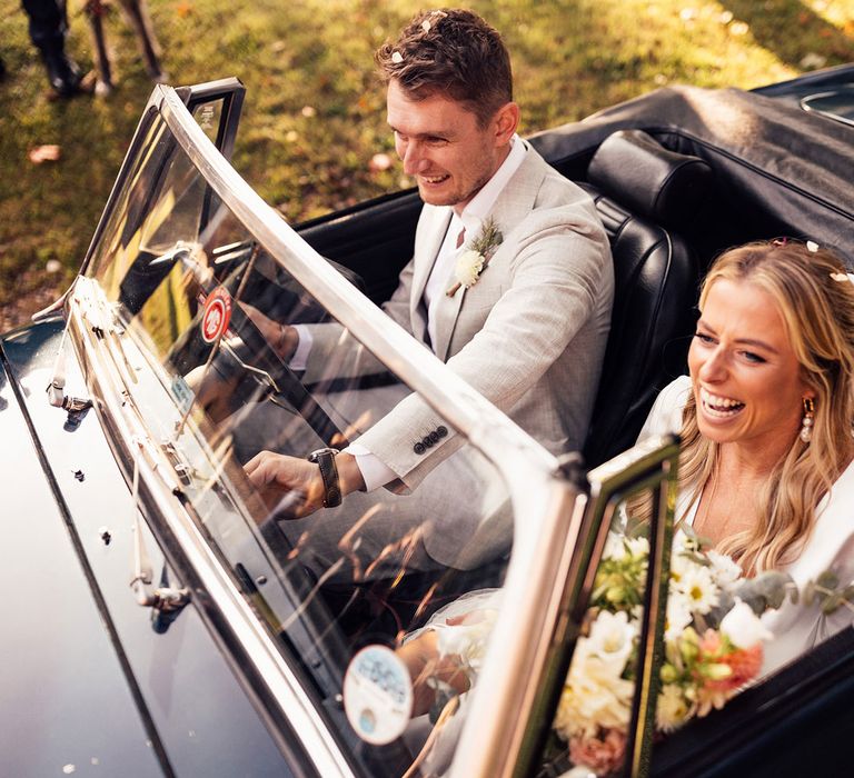 Laughing bride in hair bow and satin top holding bouquet sits in convertible car with groom in grey suit and buttonhole after wedding ceremony