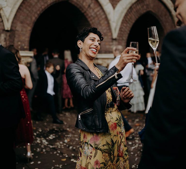 Wedding guest wears leather jacket and brightly coloured dress whilst hold champagne glass up to friend | Irene Yap Photography