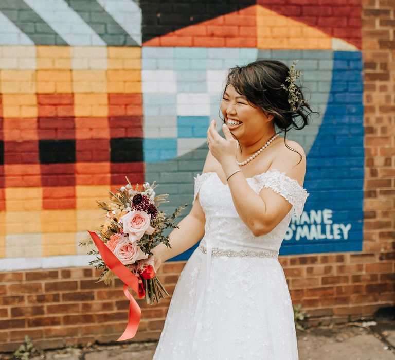 Laughing bride in wedding dress with belt holding mixed wedding bouquet with light pink roses as she stands by bright wall mural in Liverpool