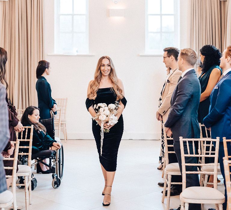 Bridesmaid in a black strapless wedding dress holding a white wedding bouquet walking down the aisle at Iscoyd Park 