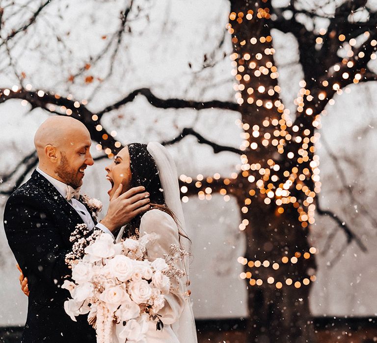 Groom in a black suit kissing his bride Jessamy Stoddart at their Iscoyd Park wedding with snow, fairy lights and white wedding flowers