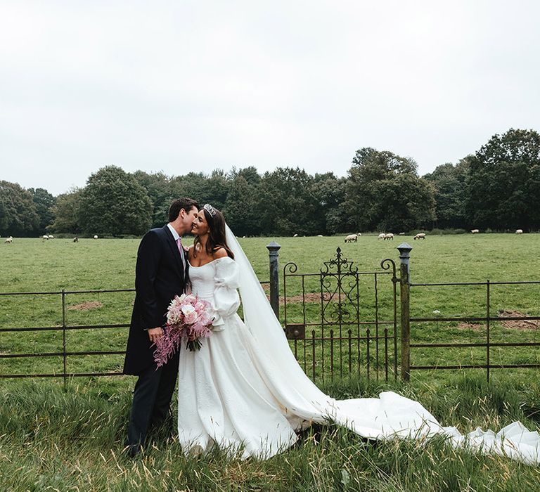 Groom in black suit and pink tie with bride in Emma Beaumont dress with long veil and tiara crown at English countryside