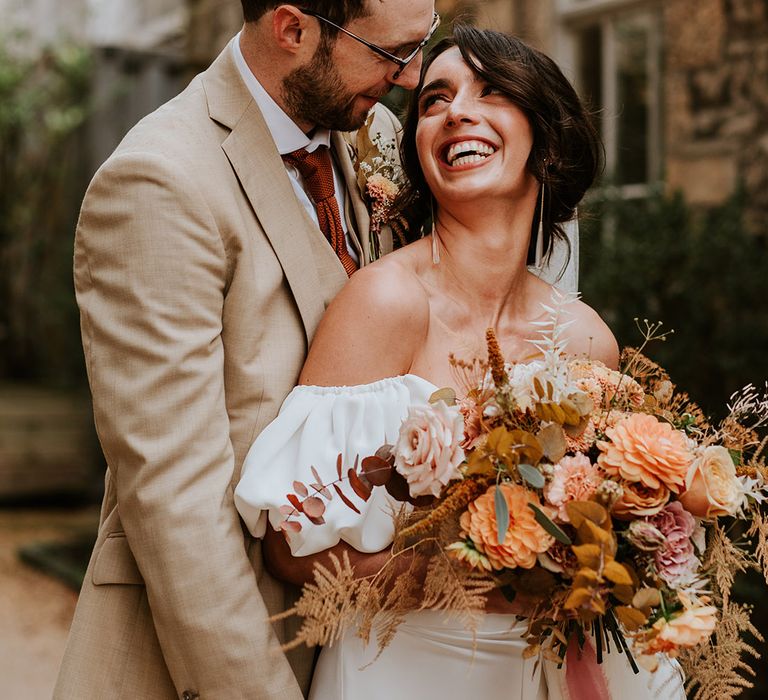 Groom in beige suit with orange tie hugs his bride form behind with bride holding autumnal wedding bouquet 