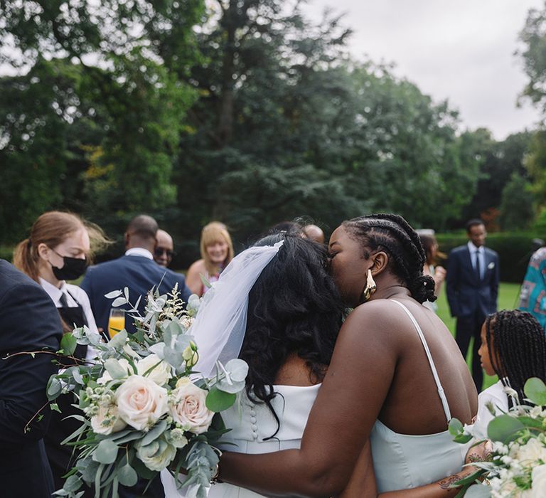 Bridesmaid in mint green dress kisses bride with white flower and green foliage bouquet