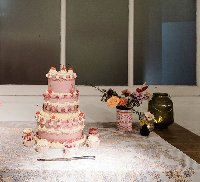 Three tier purple and white wedding cake with cherries and cupcakes in the same style as well as flowers in tin can and coloured glass vases