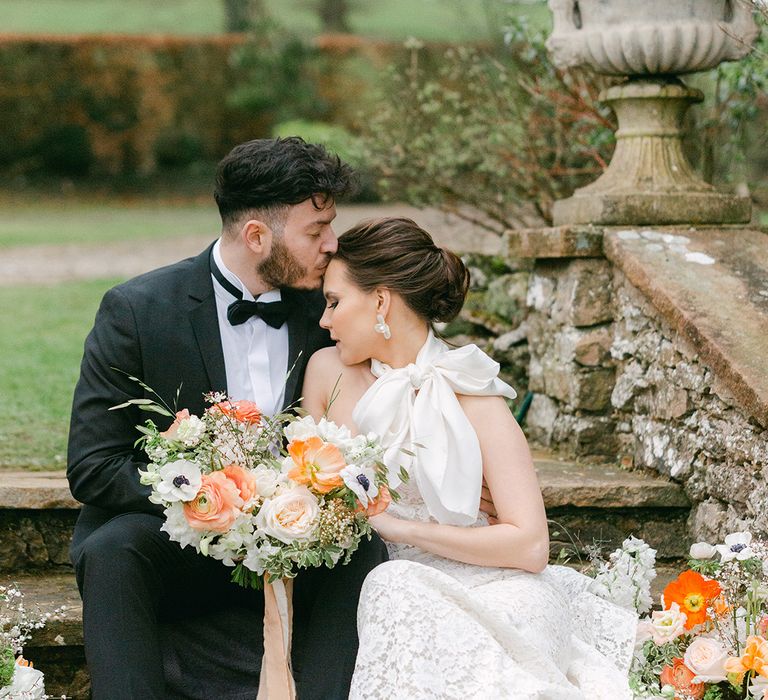 Groom in a tuxedo kissing his brides head in a lace wedding dress with bow shoulder detail holding a peach and white wedding bouquet 