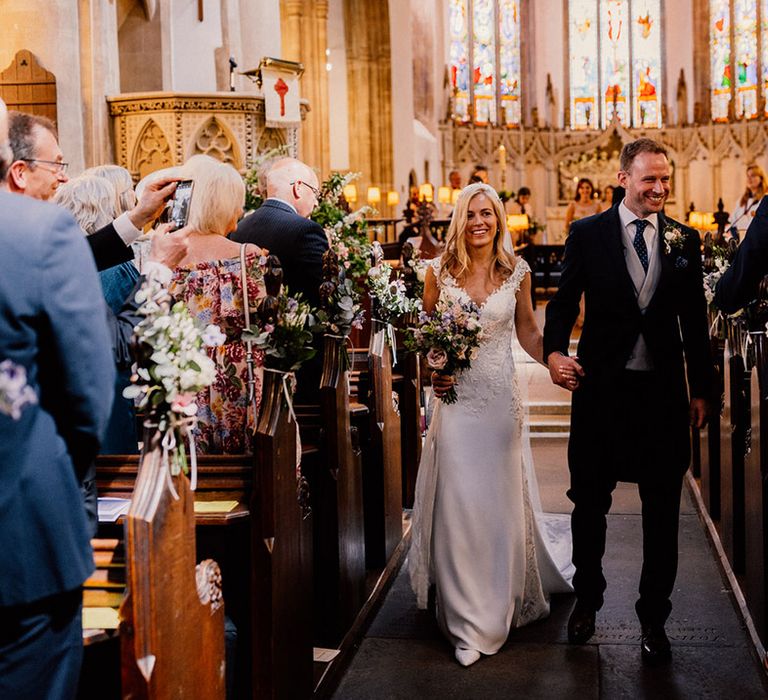 Bride in lace wedding dress walks back down the aisle with groom in morning suit with grey waistcoat as they are a married couple 