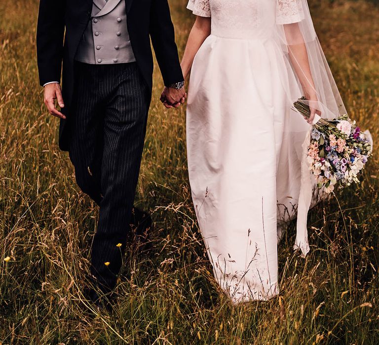 Bride and groom walk through a field holding hands as they smile at each other 