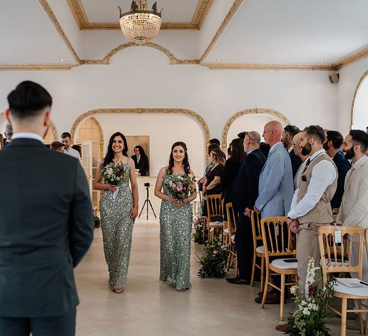 Bridesmaids wearing fitted sequin green bridesmaids dresses whilst carrying meadow floral bouquets walk down the aisle