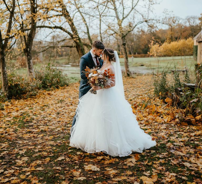 Bride and groom lean in close together as they stand in amongst autumn leaves 