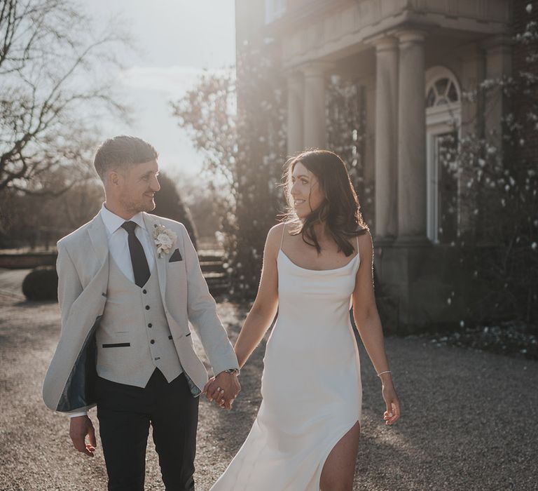 Bride in satin wedding dress with front slit and groom in grey suit jacket walk holding hands 