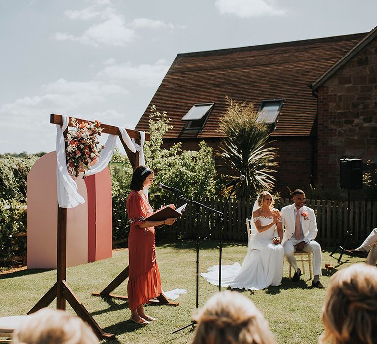 Celebrant standing in front of DIY wooden arch with drapes and pink flowers for outdoor ceremony 