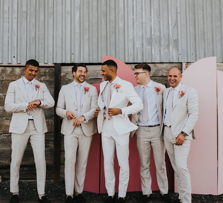 Groom in a white suit and groomsmen in cream suits with pink ties and pink flower buttonholes in front of the pink wedding screen background