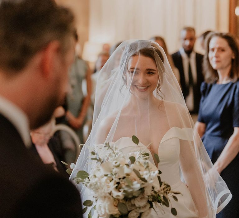 Bride in an off the shoulder wedding dress walks down the aisle to meet the groom with her veil over her face 