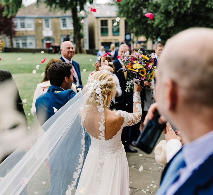 Bride & groom leave church ceremony as brides lace edged veil blows in the wind 