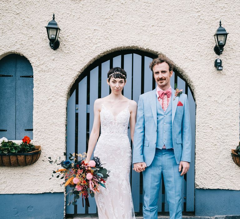 Bride wearing lace Enzoani wedding dress and holding colourful dried floral bouquet stands beside her groom in dusty blue suit and pink shirt and bow tie