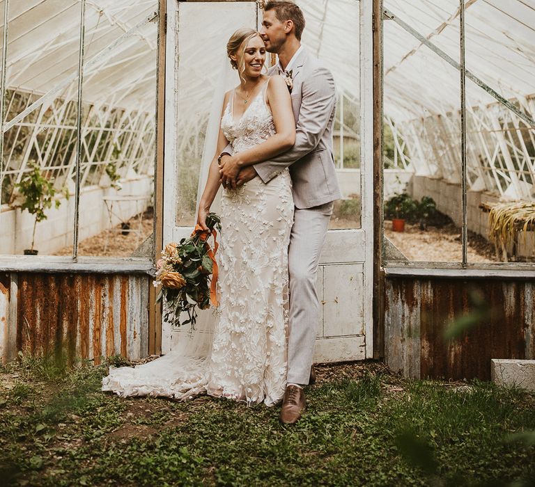 Groom in a light suit embraces the bride from behind in a lace wedding dress in front of a greenhouse at Nancarrow Farm 