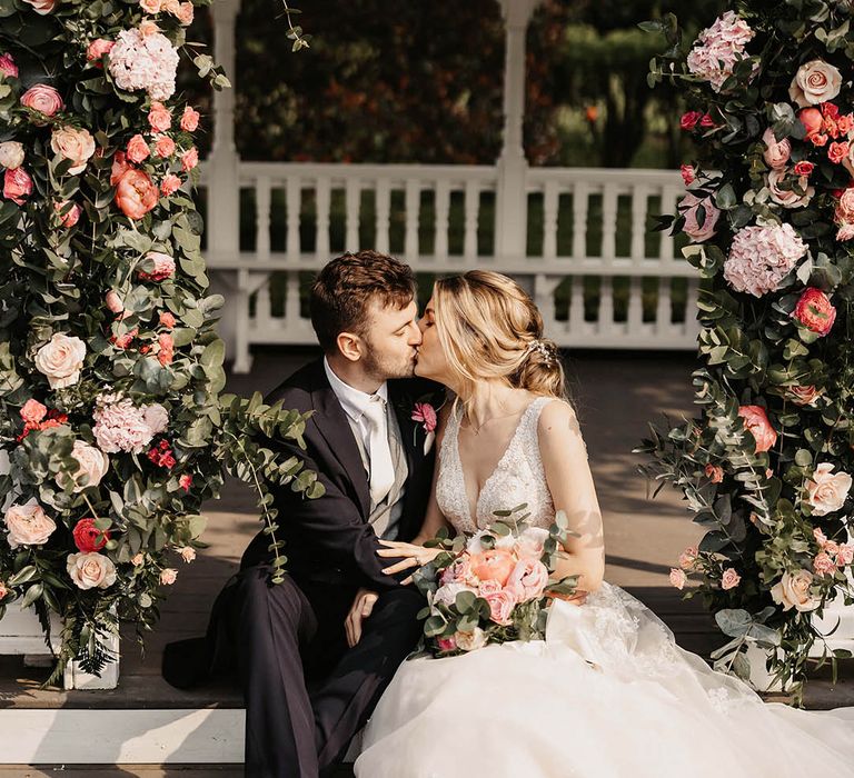 Bride and groom sit on the steps and share a kiss with a pink flower arch 