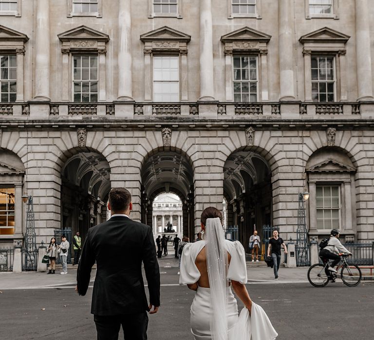 Bride in puff sleeve open back wedding dress with pearl detailed lace veil and groom in dark tux walking grounds of Somerset House