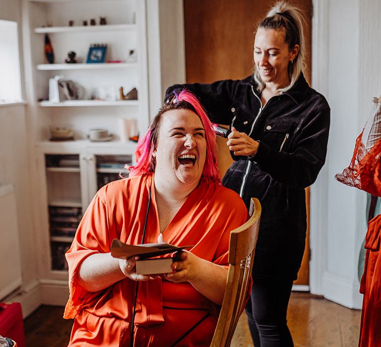 Bride in satin orange wrap around robe getting hair and makeup done with bridesmaid in orange lace wrap around robe