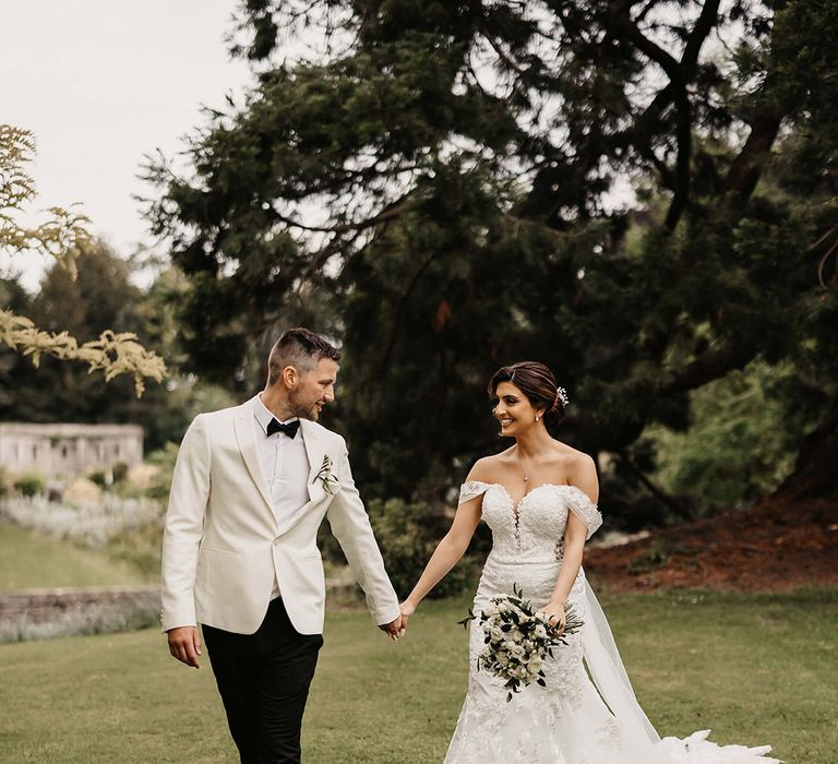 The groom in a white tuxedo walks hand in hand with the bride in a flower lace wedding dress and white flower bouquet 