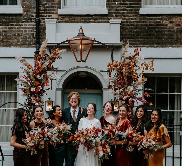 Bride in long sleeve lace vintage wedding dress and closed toe satin heels, groom in dark suit with orange tie and autumnal boutonniere and bridal party in mismatched velvet bridesmaid dresses all holding large autumnal bouquets, standing underneath large floral arch with roses, peonies, foliage and pampas grass at The Zetter Townhouse 