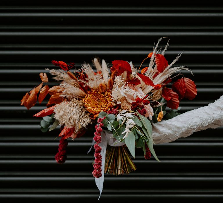 Bride holding large autumnal bouquet with orange, black, green and yellow roses, peonies, foliage and pampas grass