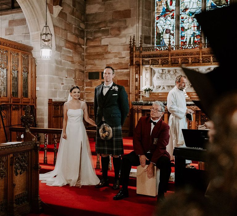 Bride in sparkly wedding dress and groom in kilt stand as the choir plays 