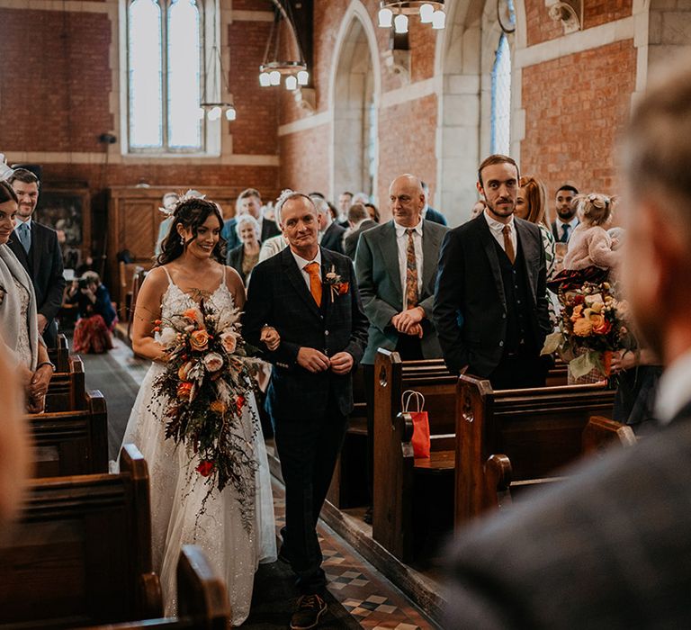 Bride in lace wedding dress holding a large cascade wedding bouquet walks down the aisle with the father of the bride in a navy suit, orange tie and pocket square 