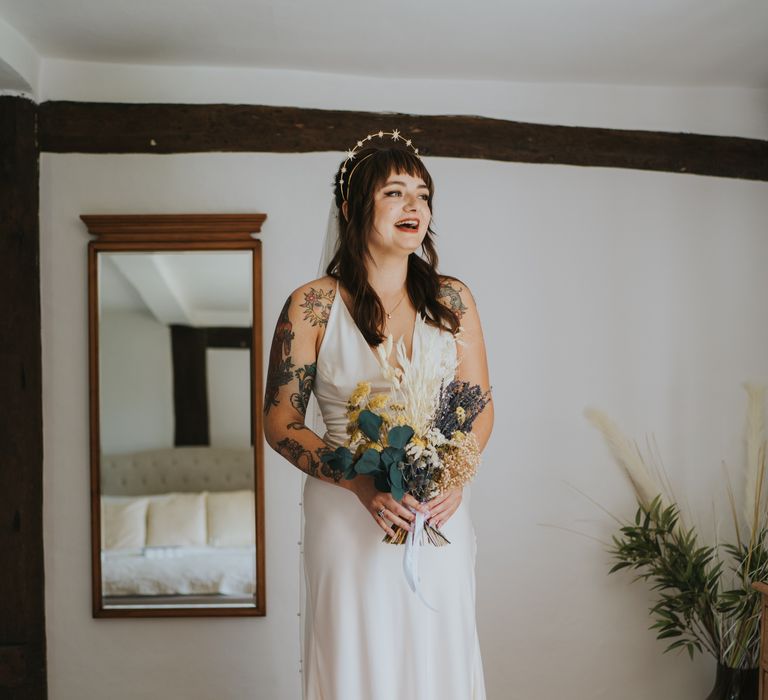 Bride getting ready for wedding, laughing, holding a dried flower wedding bouquet with white bunny tails, green foliage, yellow dried gypsophila, lavender and baby’s-breath wearing ASOS v-neck wedding dress