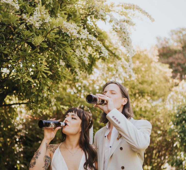 Bride in silk-look v-neck sleeveless ASOS wedding dress and gold halo headband with sparkly stars and groom in neutral coloured double-breasted ASOS suit and black high-top converse with dried flower and foliage boutonniere drinking beer after wedding