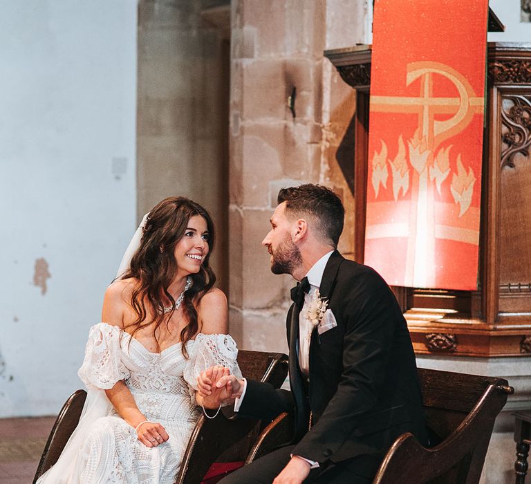 Groom in a black tuxedo with dried flower buttonhole sitting with the bride for the church ceremony in a lace wedding dress with detachable sleeves 